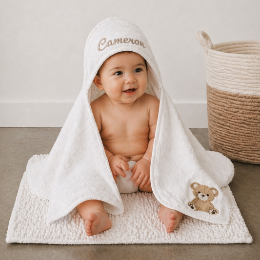 Baby sitting under a white hooded towel with 'Cameron' embroidered on it, on a neutral background.