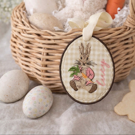 Wicker basket with plush bunny, Easter eggs, and carrot, featuring a decorative tag with rabbit illustration.