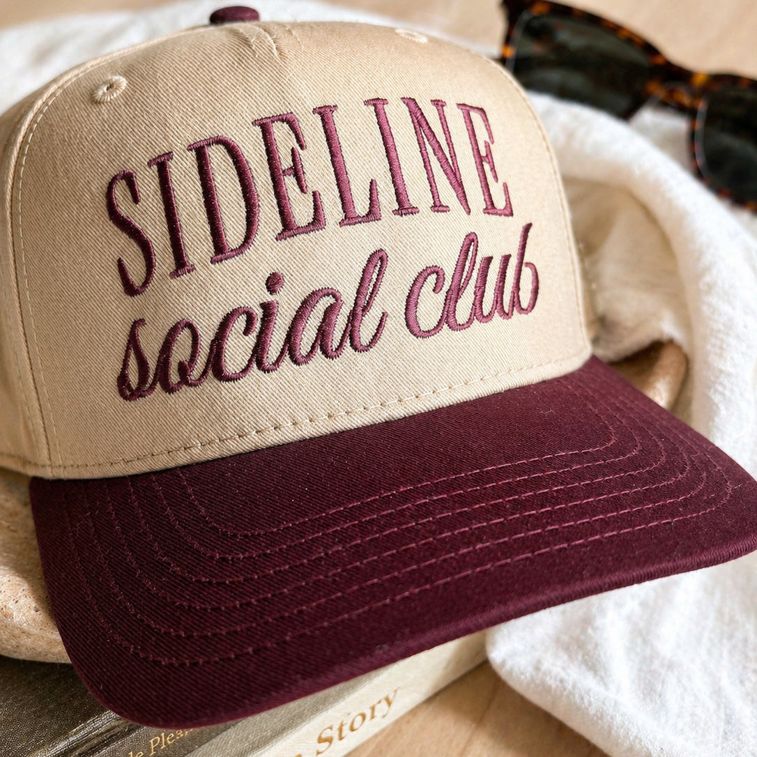 Beige and burgundy cap with 'Sideline Social Club' text on a wooden surface.