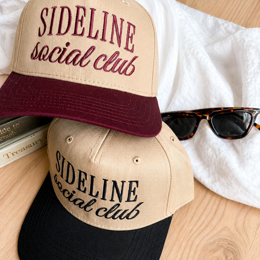 Two baseball caps with 'Sideline Social Club' text on a wooden surface.